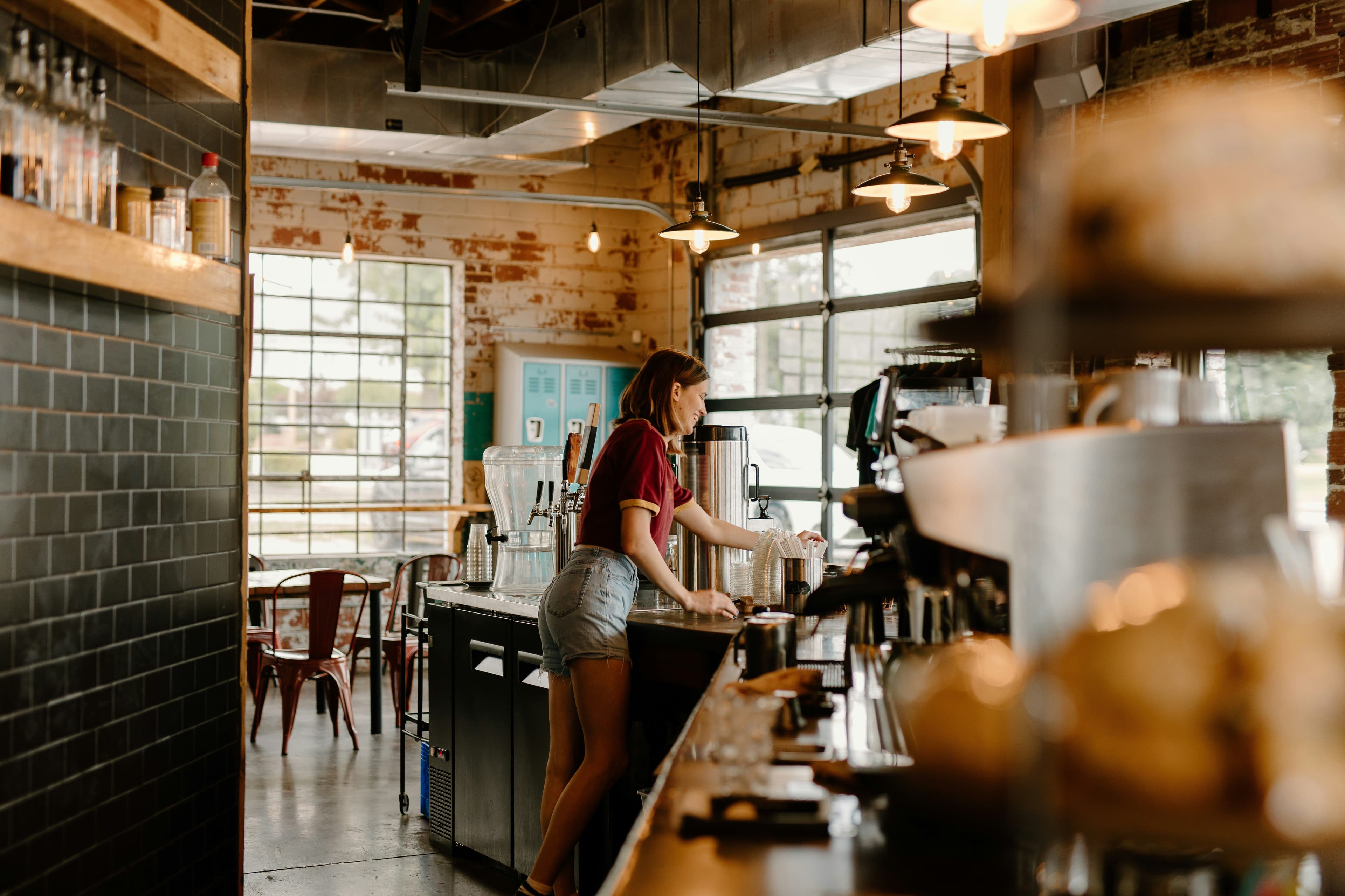 Jeune femme préparant une boisson derrière le comptoir d’un café au décor industriel avec murs en briques apparentes.