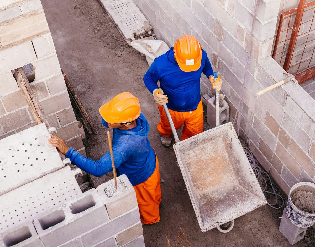 Deux ouvriers en casque orange et vêtements de chantier manipulant une brouette entre des murs en parpaings.