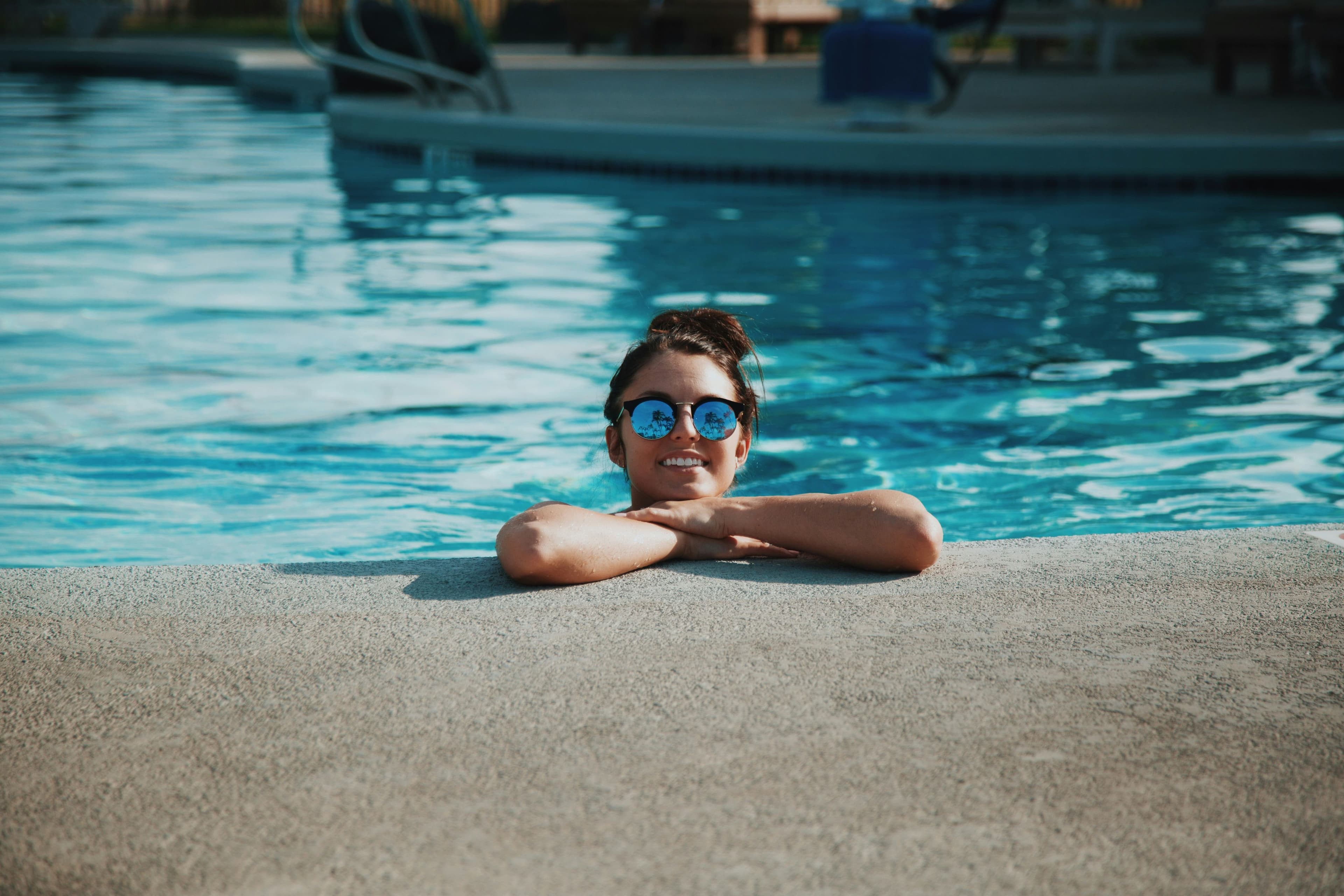 Femme souriante avec lunettes de soleil appuyée sur le bord d'une piscine, partiellement immergée dans l'eau bleue.
