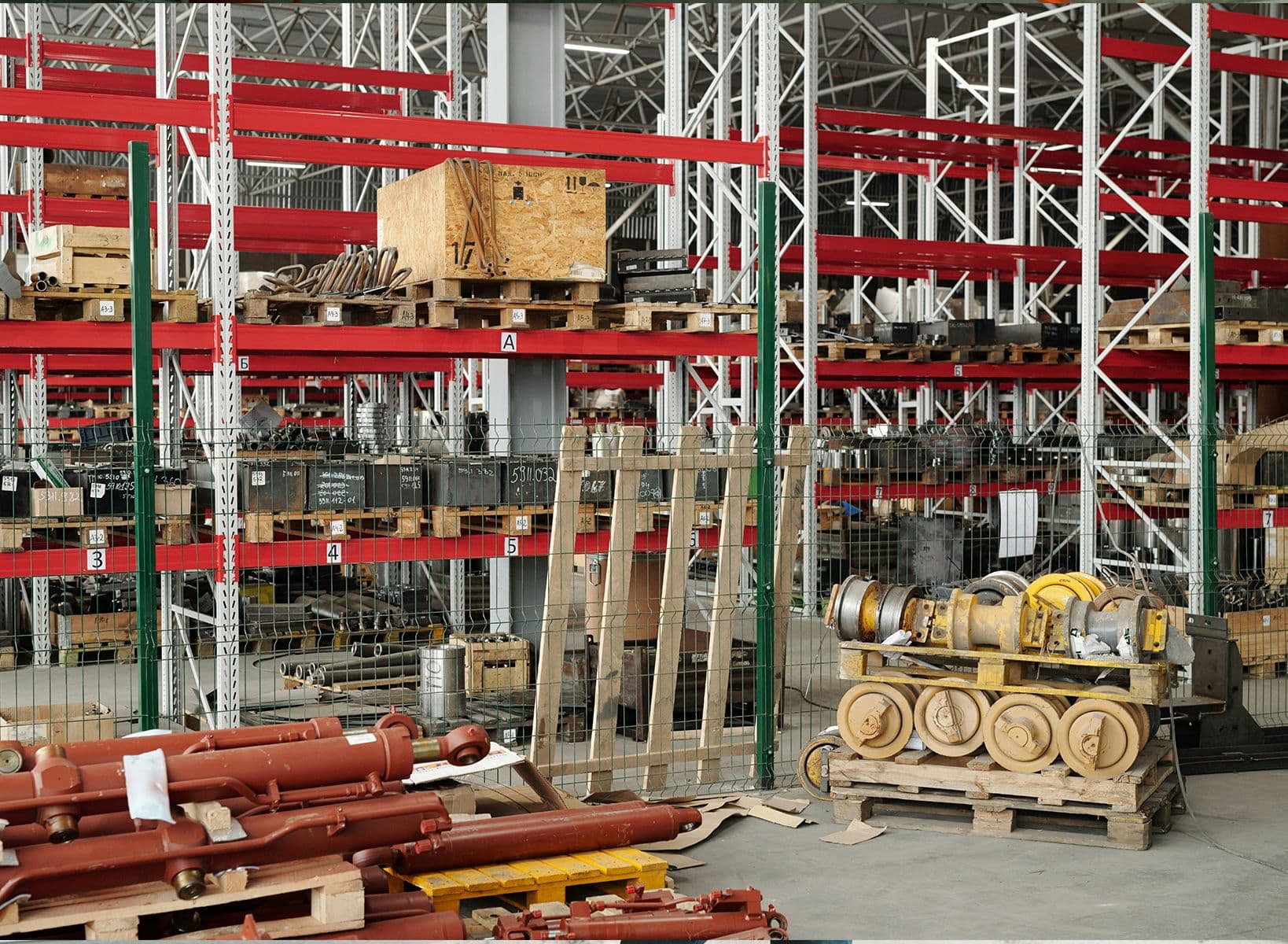 Entrepôt industriel avec étagères métalliques rouges remplies de pièces mécaniques, palettes et caisses en bois.