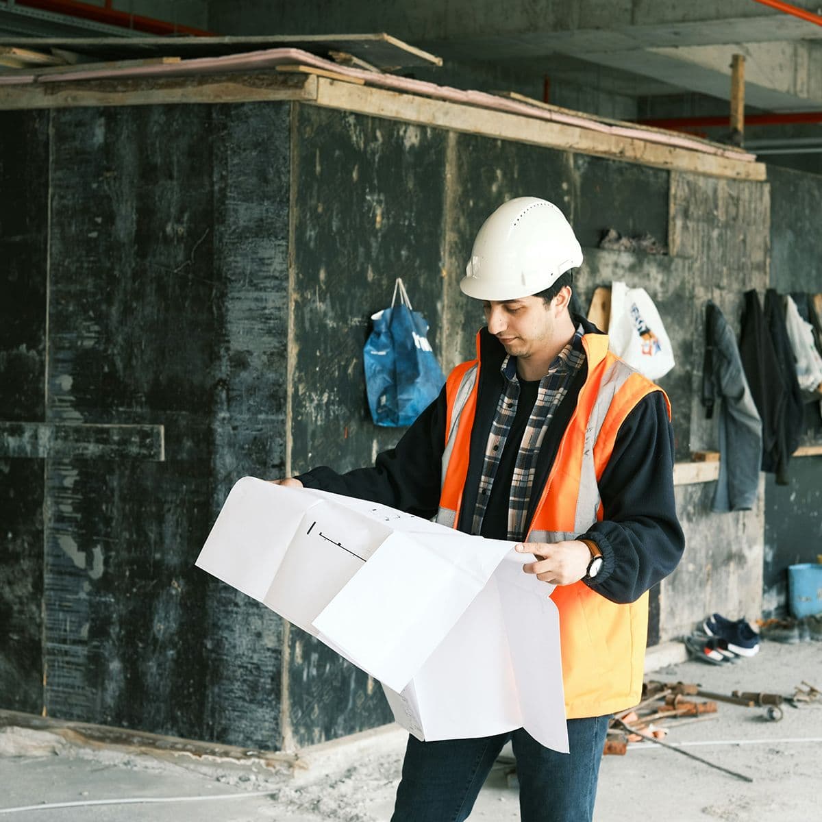 Homme en gilet orange et casque blanc consultant un plan de construction sur un chantier en intérieur.