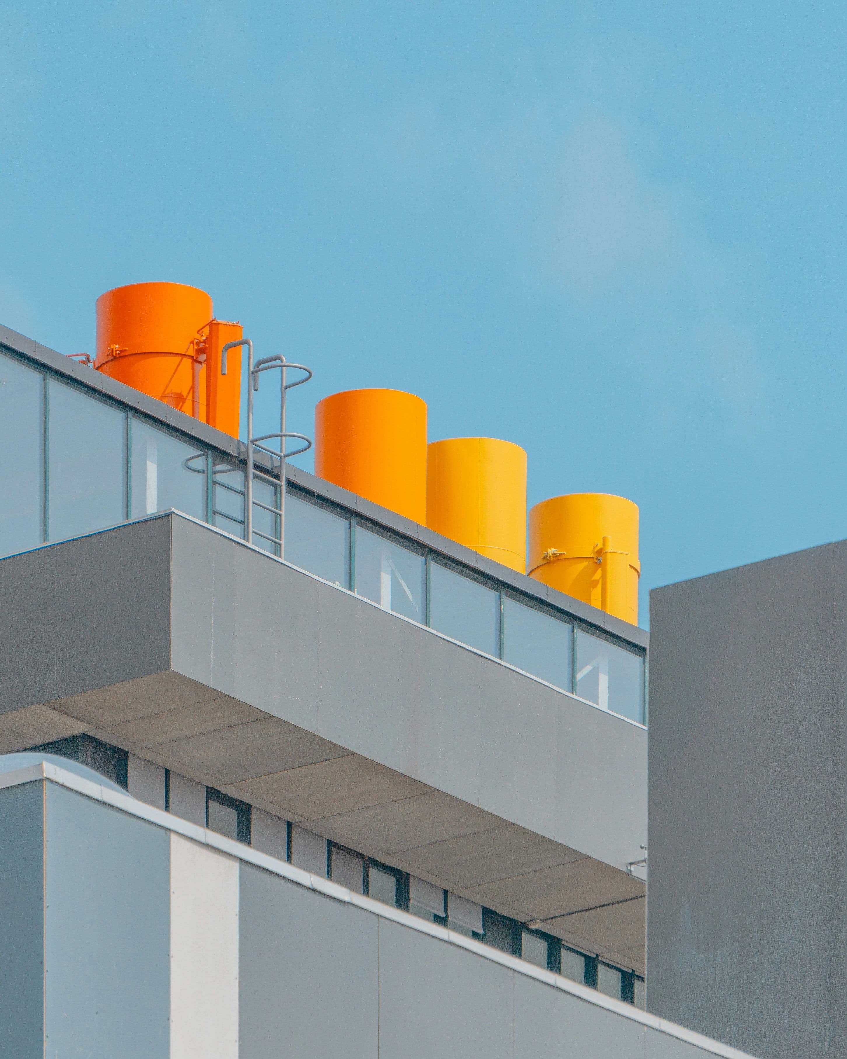Toit d’un bâtiment moderne avec quatre cheminées cylindriques colorées en orange et jaune sous un ciel bleu.