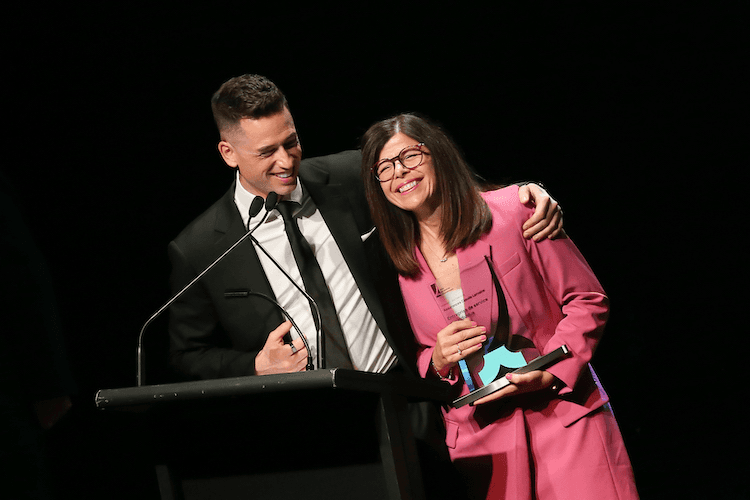 Vincent Lemoine serrant l’épaule d’une femme en tailleur rose tenant un trophée, sur une scène avec un podium.