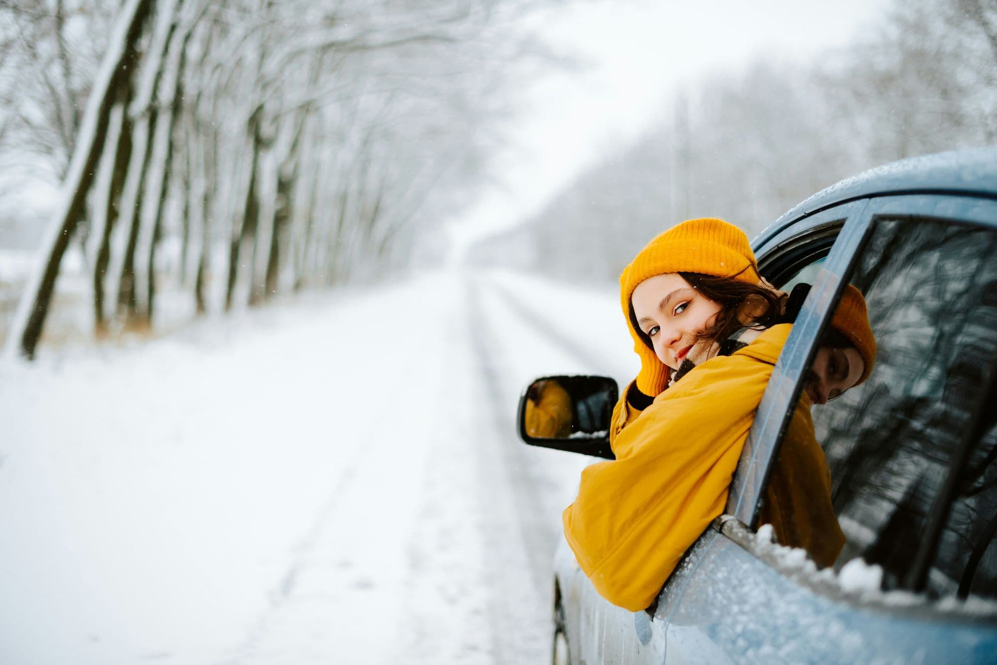 Jeune femme en bonnet orange regardant par la fenêtre d'une voiture sur une route enneigée bordée d'arbres.