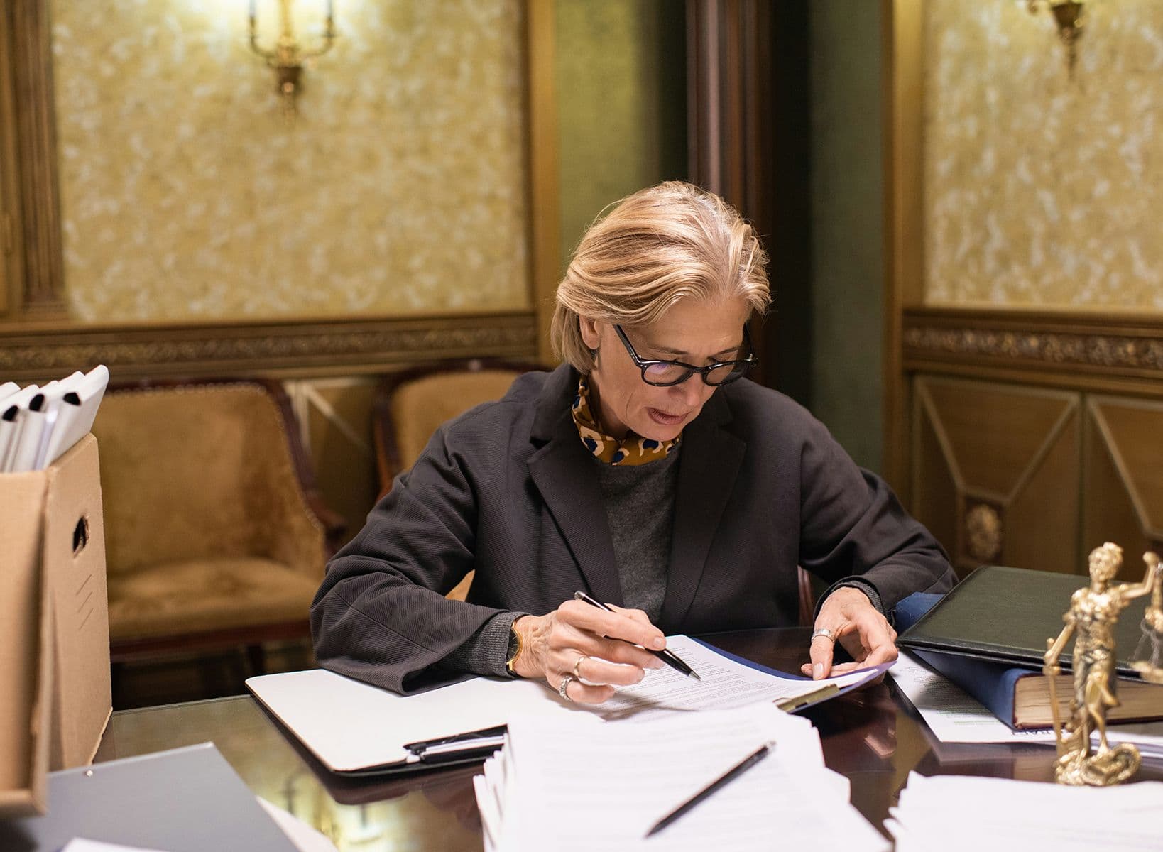 Femme concentrée en tenue professionnelle travaillant sur des documents à un bureau, dans un cabinet au décor classique.