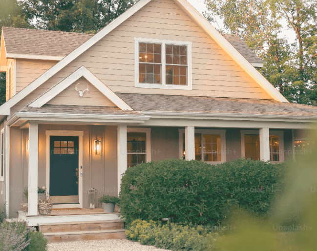 Maison familiale beige avec porche, porte d’entrée sombre et jardin soigné, éclairée par une lumière douce en fin de journée.