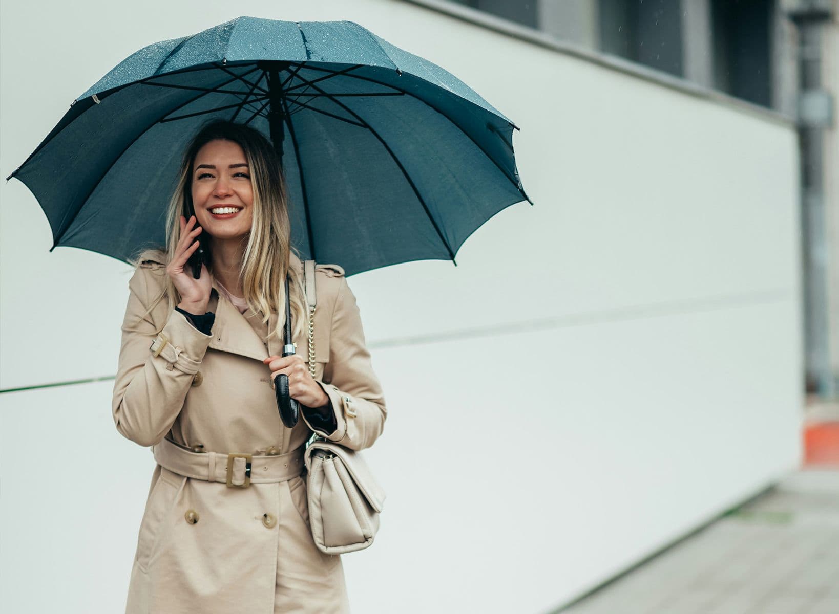 Femme souriante en imperméable beige parlant au téléphone sous un parapluie bleu par temps de pluie.